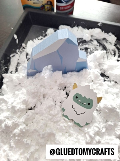 A paper yeti and an igloo-shaped cutout sit on a tray of fake snow, with the Instagram handle @GLUEDTOMYCRAFTS visible in the foreground.