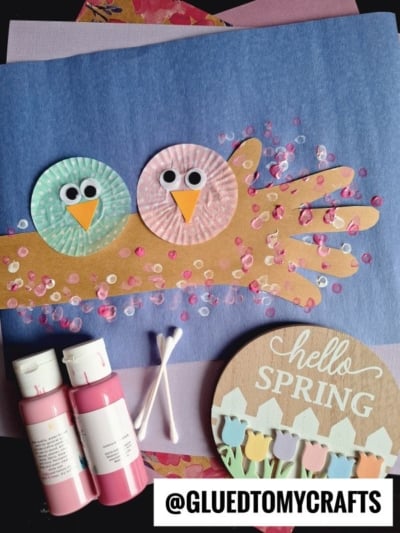 A spring craft featuring two birds made from pastel cupcake liners and googly eyes sitting on a cut-out paper hand branch, with pink blossoms made from paint and cotton swabs—perfect for creative cupcake liner card crafts. Hello Spring sign and supplies nearby.