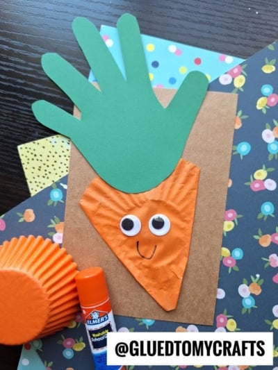 A fun cupcake liner card craft: a carrot made with a green handprint top, orange cupcake liner body, googly eyes, and a drawn smile on floral-patterned paper—perfect for creative kids! @GLUEDTOMYCRAFTS label at the bottom.