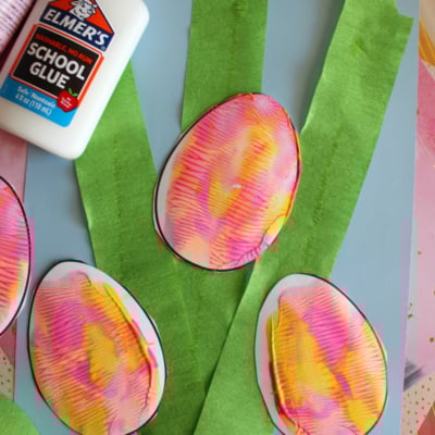 Several colorful, paint-splattered paper eggs are displayed on a blue background with green tissue paper strips resembling grass. A bottle of Elmers glue and the website “GluedToMyCraftsBlog.com” are visible. Text reads Paint Splat Eggs.