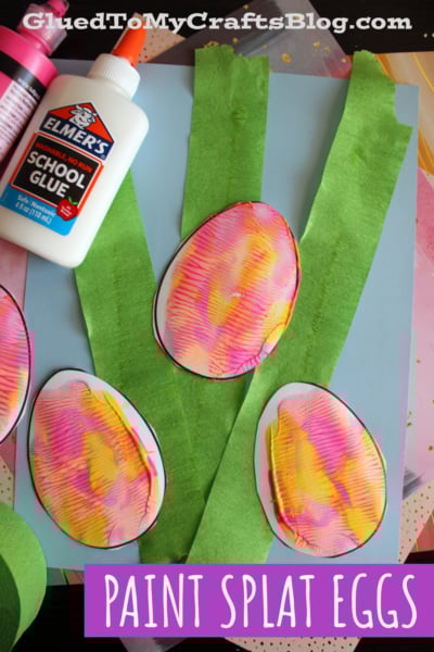 Several colorful, paint-splattered paper eggs are displayed on a blue background with green tissue paper strips resembling grass. A bottle of Elmers glue and the website “GluedToMyCraftsBlog.com” are visible. Text reads Paint Splat Eggs.