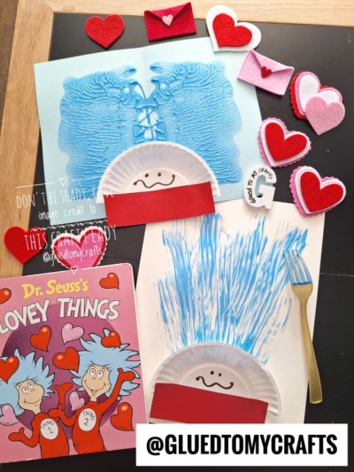 A flat lay of Valentines crafts: two blue painted paper plate faces with red paper headbands, felt hearts, mini envelopes, a Dr. Seusss Lovey Things book, and a fork with blue paint on a chalkboard surface.