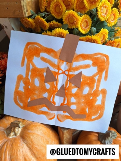 A white paper with a symmetrical orange paint design resembling a pumpkin, decorated with brown paper eyes, nose, and mouth. Surrounded by orange flowers and pumpkins—perfect inspiration for FALL CRAFTS FOR KIDS. @GLUEDTOMYCRAFTS text at the bottom.