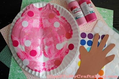 A pink painted paper plate mask with colorful dot stickers, surrounded by a paper hand cutout, dot sticker sheets, bottles of pink paint, and patterned craft paper.