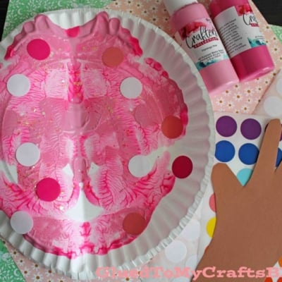 A pink painted paper plate mask with colorful dot stickers, surrounded by a paper hand cutout, dot sticker sheets, bottles of pink paint, and patterned craft paper.