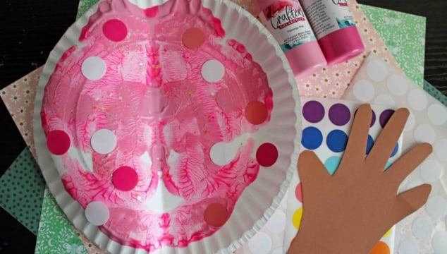 A pink painted paper plate mask with colorful dot stickers, surrounded by a paper hand cutout, dot sticker sheets, bottles of pink paint, and patterned craft paper.
