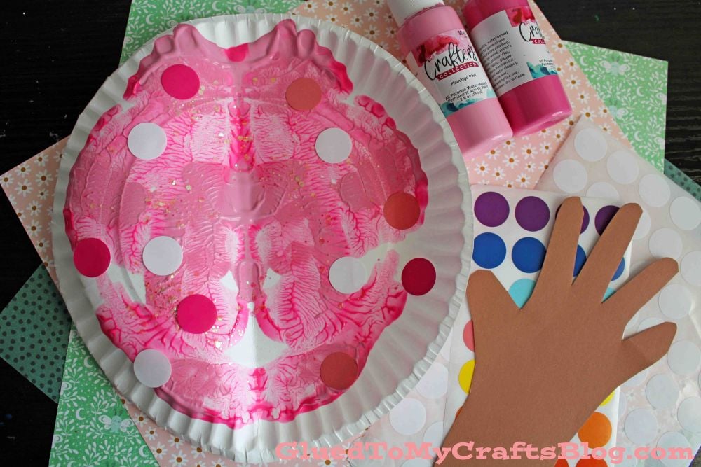 A pink painted paper plate mask with colorful dot stickers, surrounded by a paper hand cutout, dot sticker sheets, bottles of pink paint, and patterned craft paper.