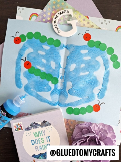 A blue butterfly painting on paper, decorated with a green caterpillar made from paper circles and red dots for the head. Craft supplies and a children’s book titled “Why Does It Rain?” are nearby. The handle reads “GLUED TO MY CRAFTS.”.