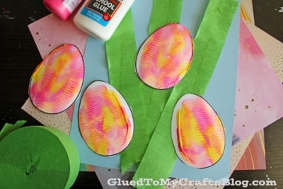 Colorful paper eggs with pink and yellow patterns are placed on top of green crepe paper strips, blue paper, and other craft materials. A bottle of school glue is in the upper corner.
