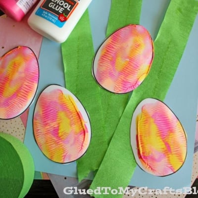 Colorful paper eggs with pink and yellow patterns are placed on top of green crepe paper strips, blue paper, and other craft materials. A bottle of school glue is in the upper corner.