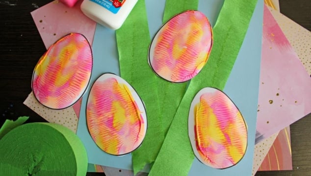 Colorful paper eggs with pink and yellow patterns are placed on top of green crepe paper strips, blue paper, and other craft materials. A bottle of school glue is in the upper corner.