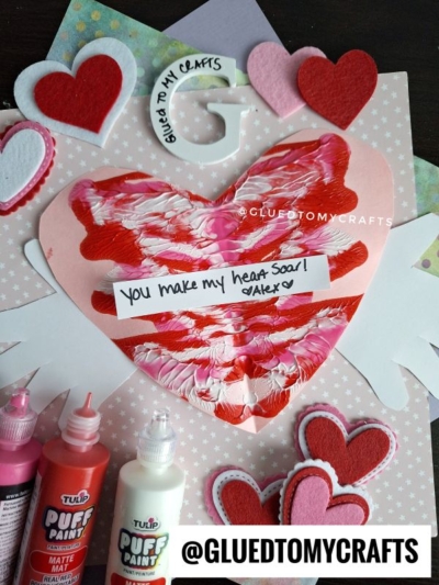 A handmade Valentine’s card features a large heart with red, pink, and white paint swirls, a note reading You make my heart soar! Alex, felt hearts, and bottles of puffy paint. @GLUETOMYCRAFTS is written at the bottom.