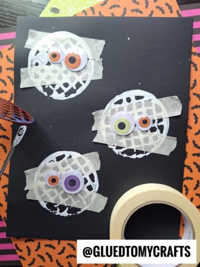 A black paper features a potato masher stamped craft with three mummy faces made from white paint circles, googly eyes, and tape strips. A roll of tape and a purple tool are nearby, set against orange and green Halloween-themed paper.