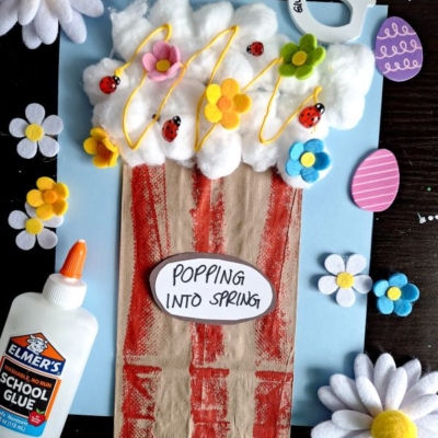 A spring-themed craft features a popcorn bucket made from brown paper and cotton balls with ladybugs, flowers, and yellow “popcorn” on a blue background. Elmer’s glue and felt flowers are nearby. Text: “POPPING INTO SPRING.”.