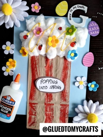 A spring-themed craft features a popcorn bucket made from brown paper and cotton balls with ladybugs, flowers, and yellow “popcorn” on a blue background. Elmer’s glue and felt flowers are nearby. Text: “POPPING INTO SPRING.”.