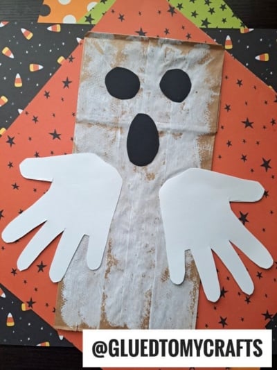 A ghost craft made from a brown paper bag painted white, with black cutouts for eyes and mouth, and two white handprint shapes as arms—perfect for Halloween Crafts for Kids on a festive holiday background.