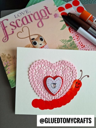 A handmade Valentine's card features a red caterpillar made from stickers on a pink heart doily—perfect inspiration for valentine's day craft ideas for kids, with markers, glue, red stickers, and the book Love, Escargot nearby.