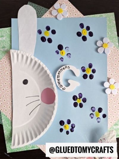 A papercraft bunny made from a white paper plate, with drawn-on face details, is displayed on pastel floral paper and blue cardstock decorated with finger-painted purple and yellow flowers and felt daisies. @GLUEDTOMYCRAFTS is written at the bottom.