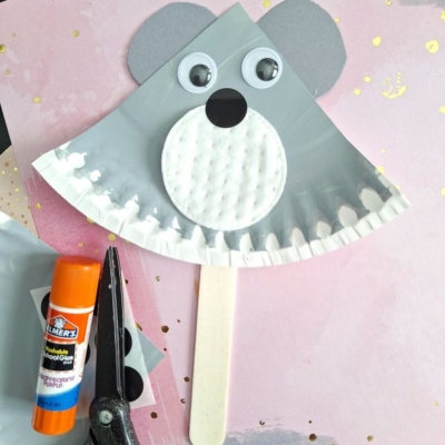 A koala craft made from a gray cupcake liner, googly eyes, paper ears, and a craft stick, lying on pink and gold paper with scissors, glue stick, and materials nearby. Text reads @GLUEDTOMYCRAFTS.