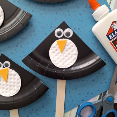 Three black paper plate penguin crafts with cotton pad bellies, googly eyes, and yellow paper beaks are displayed on a blue star-patterned background. Craft supplies like glue and scissors are nearby. @GLUEDTOMYCRAFTS is written at the bottom.