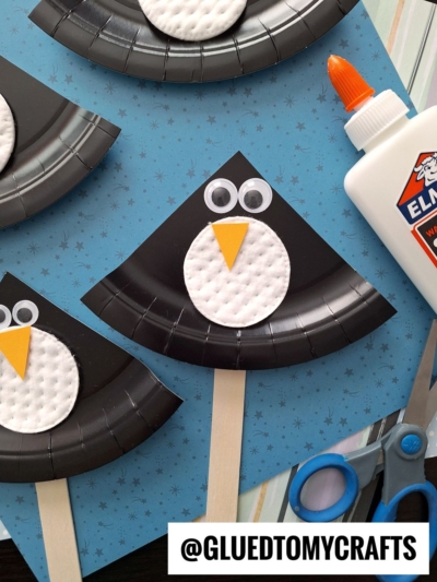 Three black paper plate penguin crafts with cotton pad bellies, googly eyes, and yellow paper beaks are displayed on a blue star-patterned background. Craft supplies like glue and scissors are nearby. @GLUEDTOMYCRAFTS is written at the bottom.