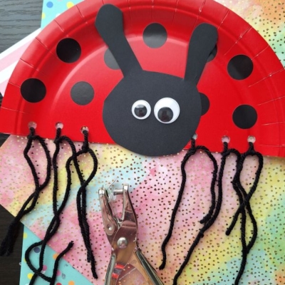A red paper plate cut in half and decorated as a ladybug with black spots, googly eyes, black antennae, and yarn legs—a fun pick for INSECT CRAFT IDEAS. A hole punch and colorful papers are nearby. A label reads @GLUEDTOMYCRAFTS.