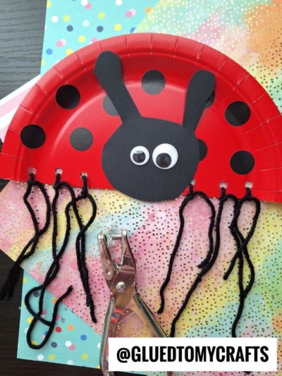 A red paper plate cut in half and decorated as a ladybug with black spots, googly eyes, black antennae, and yarn legs—a fun pick for INSECT CRAFT IDEAS. A hole punch and colorful papers are nearby. A label reads @GLUEDTOMYCRAFTS.
