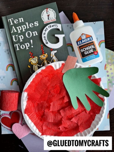A children’s book titled Ten Apples Up On Top!, a bottle of Elmer’s School Glue, red yarn, heart cutouts, and a paper plate apple craft with a green handprint leaf are on a table—perfect for Dr Seuss Kid Crafts. @GLUEDTOMYCRAFTS is at the bottom.