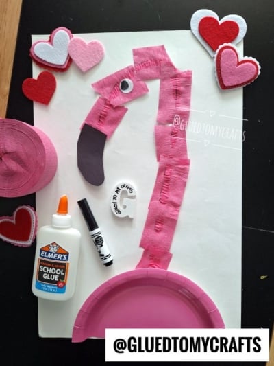 A pink flamingo craft made from a paper plate, tissue paper, felt hearts, glue, and a marker is displayed on a white background with crafting supplies around it. The @GLUEDTOMYCRAFTS watermark is visible.