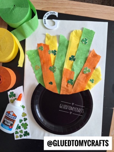 A St. Patrick’s Day craft features a black paper pot, strips of green, yellow, and orange tissue paper, shamrock stickers, a bottle of Elmer’s glue, and rolls of tissue paper on a table. @GLUEDTOMYCRAFTS is written at the bottom.
