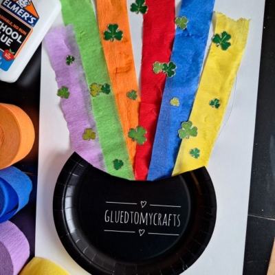 A St. Patricks Day craft features a black paper plate pot, rainbow tissue paper strips, and green shamrock cutouts. Elmers glue and rolls of colored crepe paper are nearby. Text: @GLUEDTOMYCRAFTS is visible.