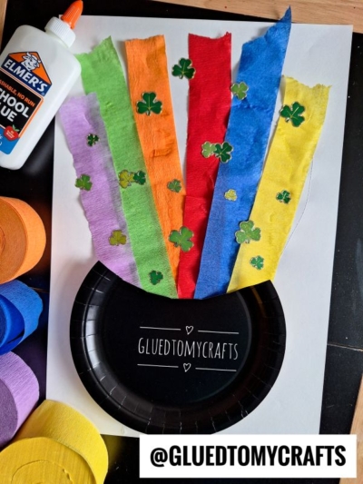A St. Patricks Day craft features a black paper plate pot, rainbow tissue paper strips, and green shamrock cutouts. Elmers glue and rolls of colored crepe paper are nearby. Text: @GLUEDTOMYCRAFTS is visible.