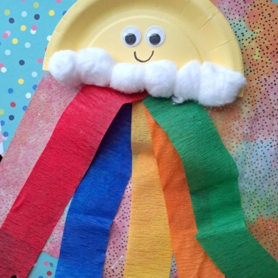 A fun crepe paper craft idea: a paper plate sun with googly eyes, cotton ball clouds, and colorful crepe paper streamers on a blue polka dot background. @GLUEDTOMYCRAFTS is written at the bottom.