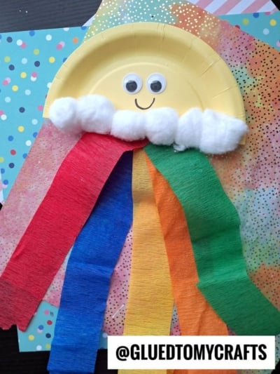 A fun crepe paper craft idea: a paper plate sun with googly eyes, cotton ball clouds, and colorful crepe paper streamers on a blue polka dot background. @GLUEDTOMYCRAFTS is written at the bottom.