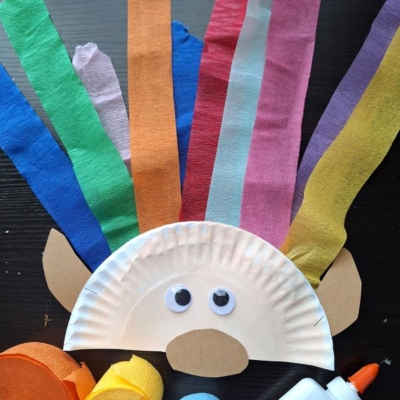 A paper plate craft resembling a face with googly eyes and brown paper ears, featuring colorful crepe paper strips for hair. Surrounded by rolls of crepe paper, glue, and the handle @GLUEDTOMYCRAFTS—perfect for crepe paper craft ideas.
