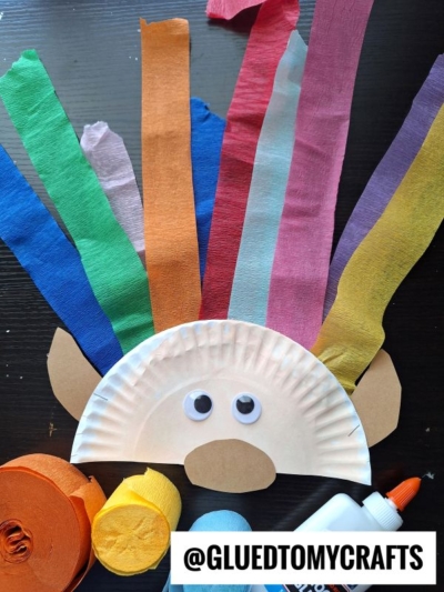 A paper plate craft resembling a face with googly eyes and brown paper ears, featuring colorful crepe paper strips for hair. Surrounded by rolls of crepe paper, glue, and the handle @GLUEDTOMYCRAFTS—perfect for crepe paper craft ideas.