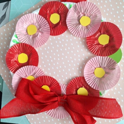 A handmade wreath made of red and pink mini cupcake liners with yellow centers, arranged in a circle on a white base. A large red ribbon is tied in a bow at the bottom. Background has patterned paper.