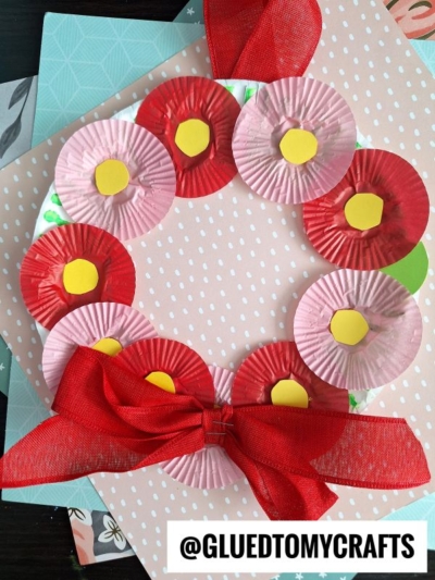A handmade wreath made of red and pink mini cupcake liners with yellow centers, arranged in a circle on a white base. A large red ribbon is tied in a bow at the bottom. Background has patterned paper.