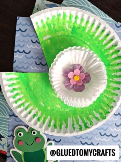 A paper plate painted green with a cupcake liner and a paper flower in the center, resembling a lily pad, sits on blue patterned paper. Perfect for cupcake liner card crafts, with a cartoon frog sticker and @GLUEDTOMYCRAFTS at the bottom.