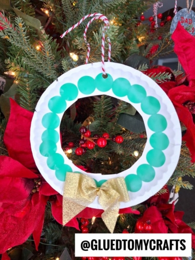A handmade wreath ornament crafted from a white paper plate with green circles, a gold bow, and a red-and-white string, hanging on a Christmas tree with poinsettias and red berries—perfect for coding label crafts this holiday season.