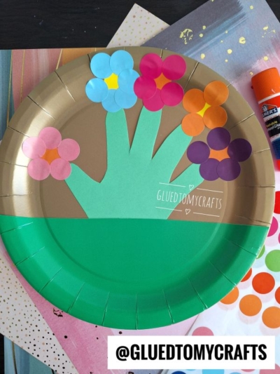A paper plate craft shows a green handprint forming the stem and leaves of a flower bouquet, with colorful paper circles as flowers. Art supplies, sticker dots, and coding label crafts are visible in the background. @GLUEDTOMYCRAFTS is written at the bottom.