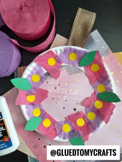 A paper plate wreath decorated with torn purple and pink crepe paper, green paper leaves, and yellow circles—a fun example of crepe paper craft ideas. Craft supplies like glue and crepe paper rolls are nearby, and the handle is made of brown ribbon.