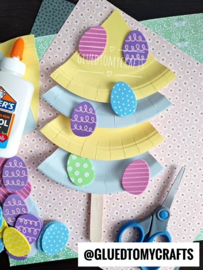 A paper Easter tree craft made from a yellow and blue paper plate, decorated with colorful paper eggs, sits on pastel background paper. Nearby are scissors, a glue bottle, and more paper eggs.
