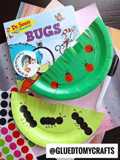 A Dr. Seuss book titled Bugs is next to two green paper plate INSECT CRAFT IDEAS: one decorated as a ladybug and the other as an ant. Colorful paper, stickers, and a black marker are also visible on the table.