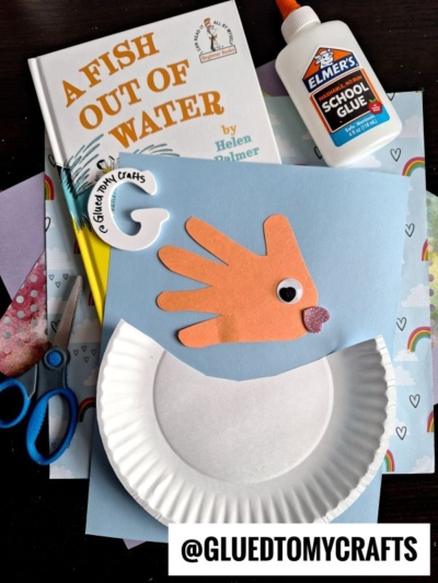 A fun Dr. Seuss Kid Crafts activity: make a fish from a blue sheet, an orange handprint, googly eye, drawn mouth, paper plate, glue, scissors, patterned papers—all inspired by the book A Fish Out of Water!.