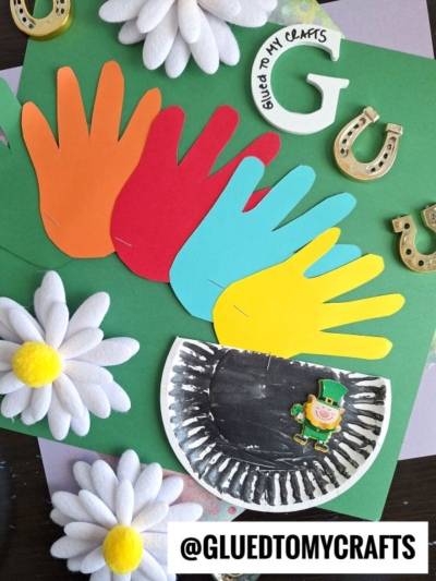 A colorful St. Patrick’s Day craft featuring four handprint cutouts (red, orange, yellow, blue), a black pot with a leprechaun sticker, felt daisies, gold horseshoes, and “@GLUEDTOMYCRAFTS” text.