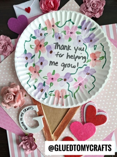 A paper plate decorated with hand-drawn flowers and the words Thank you for helping me grow! is surrounded by pink paper roses, felt hearts, and a large felt pencil cutout. The handle @GLUEDTOMYCRAFTS is at the bottom.