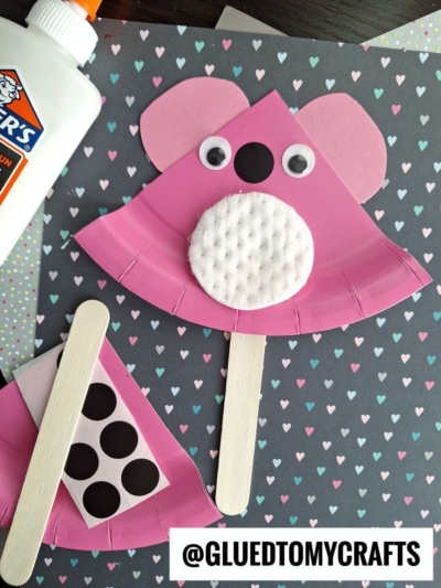 Two pink koala crafts made from paper plates, cotton pads, googly eyes, and wooden sticks are displayed on heart-patterned paper with glue, featuring the handle @gluedtomycrafts.