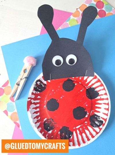 A ladybug craft made from a painted paper plate with black spots, attached to a black paper head with googly eyes and antennae. Perfect for INSECT CRAFT IDEAS, it’s paired with a clothespin topped with a pink pom-pom on colorful papers.
