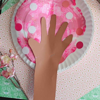 A craft project featuring a paper plate painted pink with white and pink dots to resemble cherry blossoms, and a brown paper hand cutout attached to represent a tree trunk and branches. Text reads PAINT SPLAT CHERRY BLOSSOM TREE.